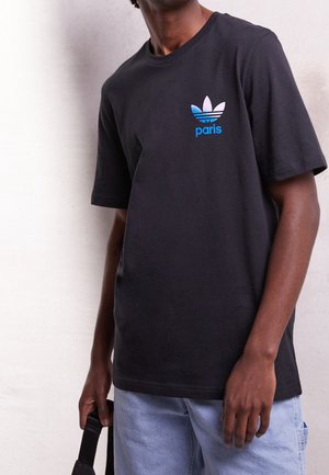 Man draagt een zwart Adidas t-shirt met blauwe "Paris"-logo, lichtblauwe jeans, en houdt een zwarte riem vast tegen een lichte achtergrond.