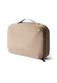 Bustina da viaggio beige realizzata in tessuto morbido con forma rettangolare. Presenta un manico superiore, chiusura con zip e una discreta etichetta del marchio sul davanti.