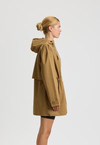 Jachetă impermeabilă maro cu glugă, talie cu snur, croială lejeră și mâneci lungi. Prezintă un design elegant și un minim de accesorii.