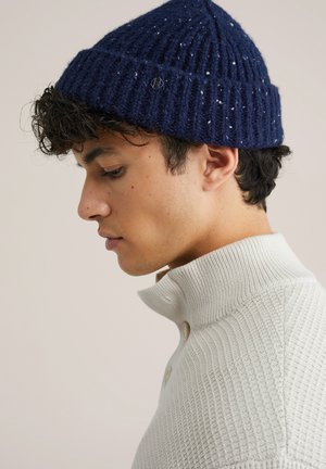 Jauns vīrietis valkā tumši zilu adītu cepuri un baltu struktūrētu džemperi ar pogām, skatoties uz leju pret vienkāršu fona.