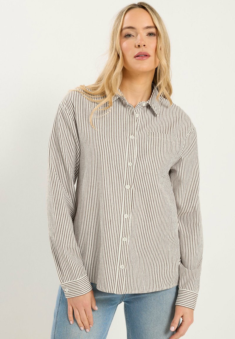 Kvinde iført en beige og sort vertikalt stribet skjorte med knapper og en forlommer samt blå jeans mod en ensfarvet baggrund.