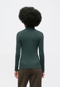 Person mit lockigem Afro, der einen dunkelgrünen, enganliegenden, langärmeligen Rollkragenpullover und braune Hosen trägt, von hinten auf einem weißen Hintergrund gezeigt.