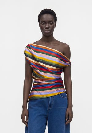Femme aux cheveux courts et bouclés porte un haut coloré rayé asymétrique, associé à un jean bleu ample sur un fond clair uni.