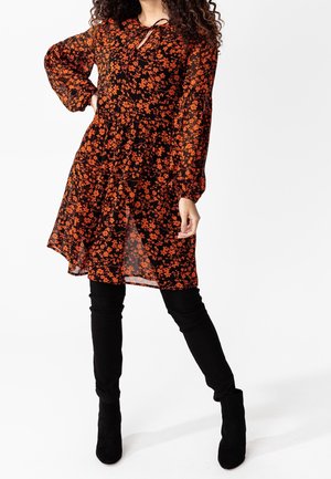 Vrouw in doorschijnende lange mouwen zwarte jurk met oranje bloemenpatroon, gecombineerd met zwarte legging en zwarte enkellaarzen, poserend tegen een witte achtergrond.