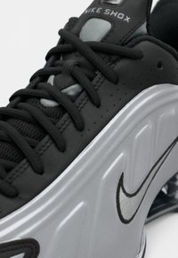 Crne Nike Shox tenisice s glatkom kožnom gornjom površinom, teksturiranim jezikom od tkanine, srebrnim potplatima i istaknutim Nike logotipom u sivoj i crnoj boji.