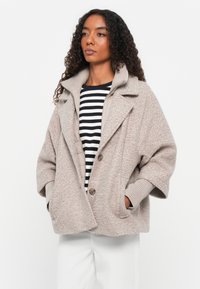 Manteau texturé beige avec un col structuré, de grandes poches avant et une fermeture à boutons, porté par-dessus un top à rayures noires et blanches.