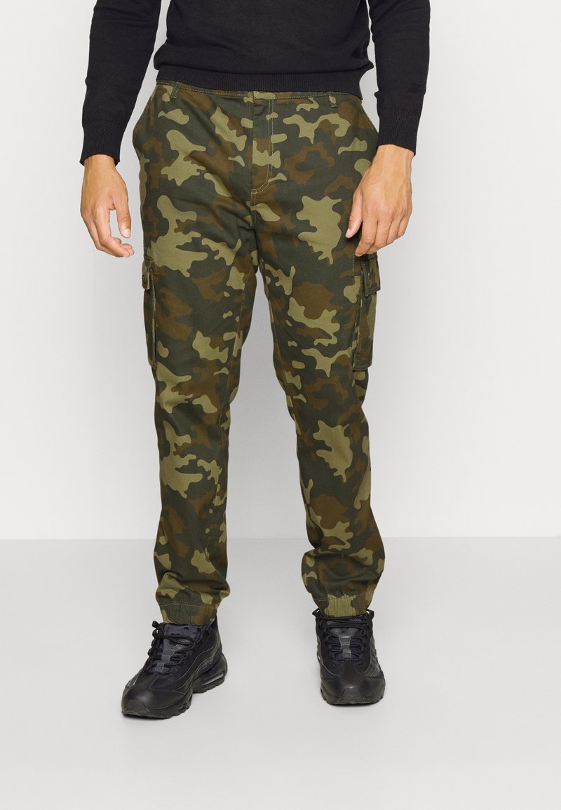 Pantalon cargo à motif camouflage avec des nuances de vert et de marron, équipé de poches latérales et de poignets élastiques, associé à des chaussures noires.