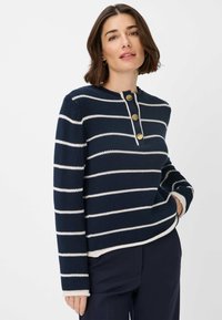 Navyblauwe gebreide trui met witte horizontale strepen, met een henley-halslijn en drie gouden knopen. Geribbelde manchetten en zoom.