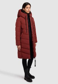Mujer vestida con un abrigo acolchado largo de color óxido, con una capucha grande, leggings negros y botas negras gruesas, de pie contra un fondo liso.