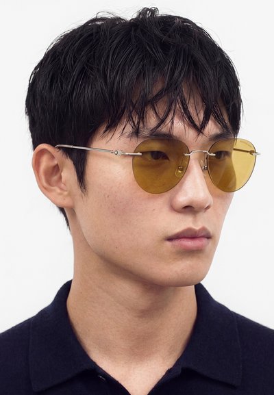 Jeune homme aux cheveux courts noirs portant des lunettes de soleil rondes teintées de jaune et une chemise sombre avec col sur fond blanc.