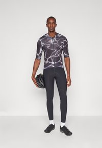 Tenue de cyclisme comprenant un maillot à manches courtes avec un motif aquatique noir et blanc, associé à un pantalon de cyclisme noir et des chaussures de cyclisme ; tenant un casque.