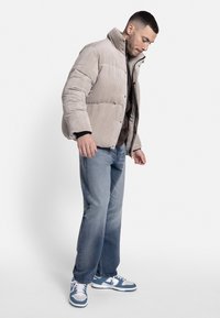 Beige corduroy pufferjack met een hoge kraag en drukknoopjes, gedragen over een bruine blouse en gecombineerd met lichtblauwe jeans en blauwe sneakers.