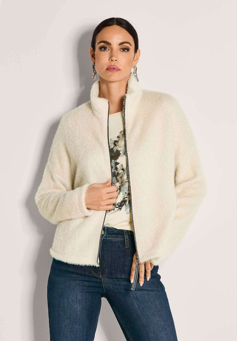 Frau in einer cremefarbenen, flauschigen Zip-Jacke, grafikbedrucktem T-Shirt, dunkelblauen Jeans und silbernen Ohrringen, die vor einem hellen Hintergrund steht.