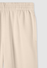 Pantaloni della tuta beige con vita elastica, texture morbida, due tasche laterali e vestibilità rilassata, mostrati su uno sfondo grigio.