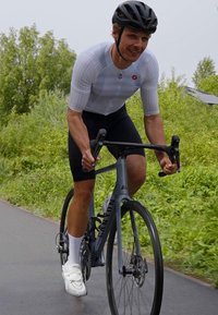 Kolarz w szarym, aerodynamicznym stroju rowerowym, jadący szarym rowerem wyścigowym z cienkimi oponami, na wybrukowanej ścieżce, otoczony zielenią.