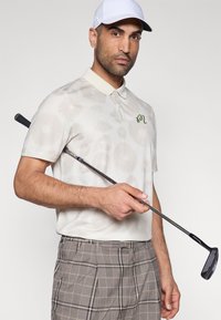 Vyriški trumpomis rankovėmis golfo polo marškinėliai smėlio spalvos su subtiliu apskritimu raštu, su kontrastine apykakle ir žaliu logotipu. Trumpi languoti golfo šortai pilki su juodais akcentais.