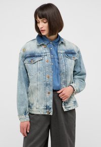 Jasnoniebieska dżinsowa kurtka o luźnym kroju, zapinana na guziki, z dwiema kieszeniami na piersi i haftowanymi akcentami. Stylizowana na niebieską koszulę chambray.