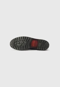 Semelle de chaussure en caoutchouc noir avec un motif de semelle profond, présentant des rainures circulaires et linéaires. Le centre a un accent rouge avec un logo embouti.