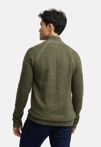 Pull en tricot vert avec un col montant et des manches raglan. Présente une surface texturée ainsi que des poignets et un ourlet côtelés.