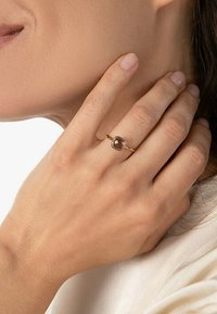 Goldring mit einem facettierten braunen Edelstein, getragen an einer Hand mit neutraler Nagelfarbe, vor einem hellen Hintergrund.