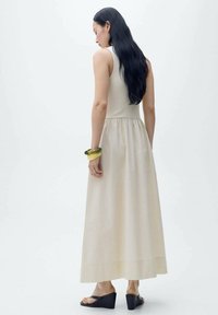 Robe maxi beige avec un corsage ajusté et une taille froncée ; sans manches. Modèle portant des sandales compensées noires et un bracelet jaune. Design simple.