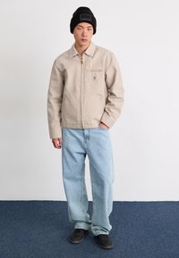 Giacca zip-up beige chiaro con taschino sul petto e logo, abbinata a jeans larghi blu chiaro e sneakers nere, in piedi su un tappeto blu.