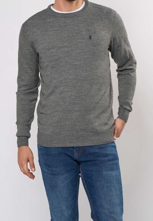 Man draagt een grijze trui met ronde hals en geribde boorden aan mouwen en zoom, gecombineerd met blauwe jeans tegen een effen witte achtergrond.