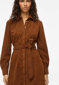 Robe chemise en velours côtelé marron avec col, manches longues, boutons sur le devant et ceinture nouée à la taille pour une silhouette cintrée.