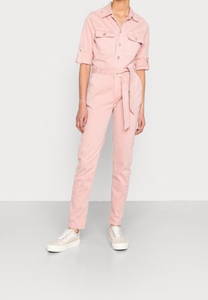 Kobieta ubrana w jasnoróżowy kombinezon z paskiem, z podwiniętymi rękawami i białe trampki, stojąca na jednolitym tle.