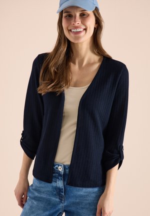 Vrouw die lacht, draagt een lichtblauwe pet, beige top, donkerblauwe openvallende cardigan met opgerolde mouwen en blauwe spijkerbroek tegen een effen achtergrond.