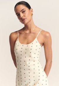 Vestido color crema hecho de tela suave, con patrones florales negros, tirantes finos y una silueta ajustada. Joyería dorada sencilla lo complementa.