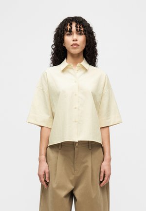 Hellgelbes Kurzarmhemd mit Knopfleiste und feiner Textur, kombiniert mit hellbraunen, zulaufenden Hosen mit Bundfalten.