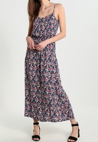 Robe longue à fleurs avec un fond bleu marine, de petites fleurs rouges et blanches, des bretelles fines réglables et une jupe fluide. Sandales à talons noires.