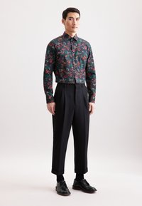 Camicia a fantasia floreale in verde scuro con accenti rossi e gialli, abbinata a pantaloni neri a vita alta e scarpe eleganti nere.