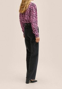 Langærmet lilla top med pink cirkulært mønster, parret med høj talje sorte jeans og sorte spidse hæle. Bagansigt.