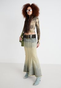 Jupe maxi en denim présentant un effet ombré allant du bleu clair au bleu foncé, associée à un haut ajusté à motifs et un col en fourrure, accessoirisée avec un sac vert.