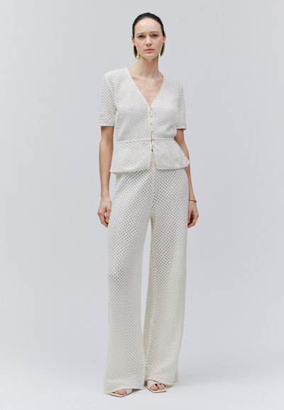 Woman with dark hair wearing white short-sleeve buttoned top and matching wide-leg pants with perforated pattern, standing against plain background.