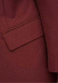 Tejido burdeos que muestra un bolsillo bien confeccionado con costuras limpias. La textura parece suave y ligeramente texturizada, enfatizando su diseño estructurado.