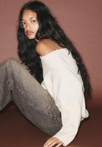 Maglione bianco a spalle scoperte abbinato a pantaloni in denim leggeri grigi con vita alta. Texture morbida, vestibilità rilassata e dettagli di cucitura ben visibili.