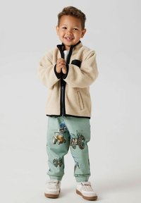 Giacca di pile beige con dettagli neri, abbinata a pantaloni verde chiaro con motivi a trattore. Sneakers bianche completano l'outfit.