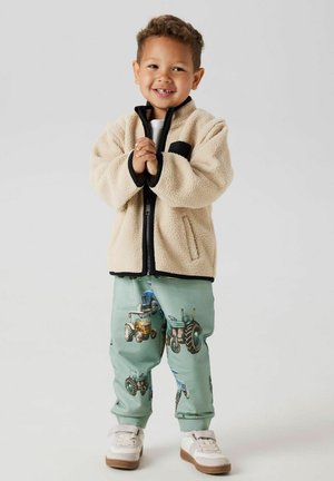 Beige fleecejack met zwarte accenten, gecombineerd met lichtgroene broeken met tractorpatronen. Witte sneakers maken de outfit compleet.