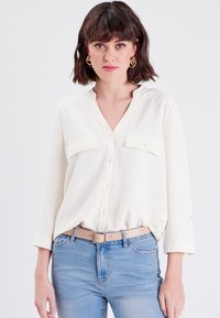 Blouse blanche à manches longues avec un col en V, deux poches avant et des boutons. Portée avec un jean bleu clair et une ceinture beige. Tissu texturé.