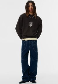 Fiatal ember hosszú fonatokkal, fekete pulóvert viselve emblémával, csíkos inget alatta, sötét bő nadrágot és fekete cipőt viselve, áll.