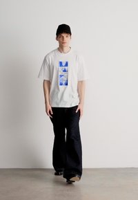 Λευκή βαμβακερή μπλούζα με μπλε γραφικά στο κέντρο, συνδυασμένη με σκούρες φαρδιές τζιν παντελόνες. Το μοντέλο φοράει ένα μαύρο καπέλο και στέκεται μπροστά σε απλό φόντο.