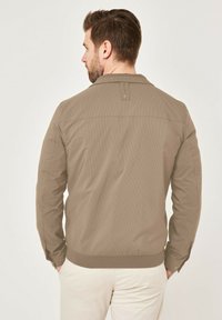 Khaki jacket with a textured, ribbed pattern. Features a collar, metal buttons, and fitted cuffs. Smooth fabric with a relaxed silhouette.