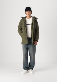 Olive green insulated jacket with hood, worn over a white ribbed sweater with navy stripes, paired with loose denim jeans and white sneakers.