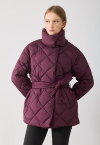 Giacca imbottita trapuntata di un profondo bordeaux con colletto alto, vita regolabile con cintura e motivo di cuciture diagonali. Texture liscia, design di lunghezza media.
