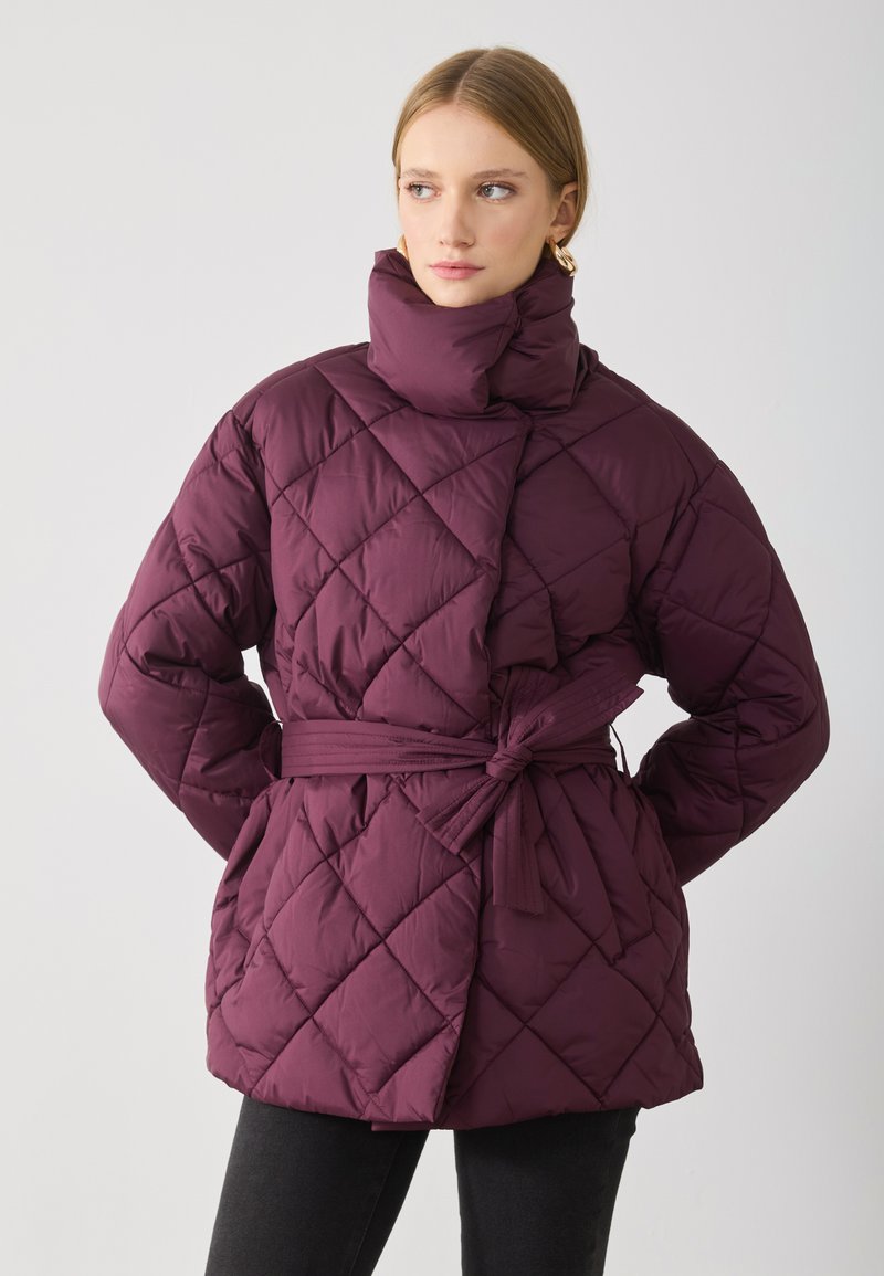 Giacca imbottita trapuntata di un profondo bordeaux con colletto alto, vita regolabile con cintura e motivo di cuciture diagonali. Texture liscia, design di lunghezza media.