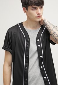Maillot noir à manches courtes avec des finitions blanches et fermeture par boutons, porté sur un t-shirt gris. Il présente un tissu texturé avec des manches retroussées.