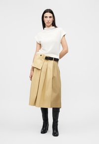 Γυναίκα που φοράει λευκό μπλουζάκι χωρίς μανίκια, μπεζ πλισέ μίντι φούστα με ζώνη-θήκη και μαύρες μπότες μέχρι το γόνατο, στέκεται μπροστά σε απλό φόντο.